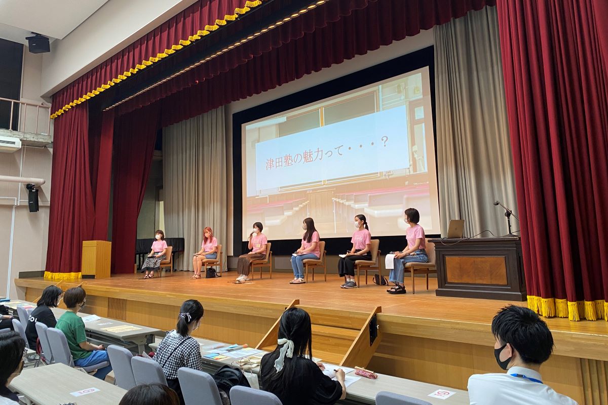 写真：壇上に6名の在学生。スクリーンには「津田塾の魅力って…？」と表示されている。