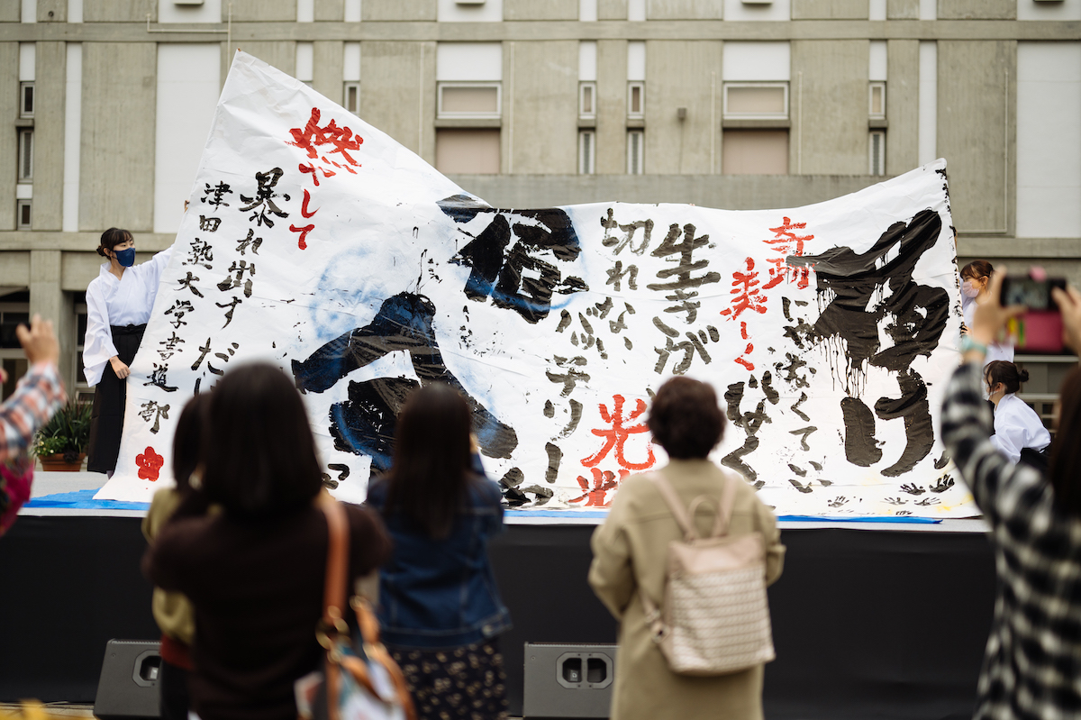 写真：中庭で、津田塾大学書道部による書道パフォーマンスが行われている。大きな紙に書かれた、「届け」「宿命」などの文字が見える。